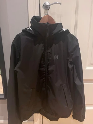 Svart vindjacka från Helly Hansen - Snygg svart vindjacka från Helly Hansen med huva och dragkedja framtill. Jackan har två sidofickor med dragkedja och HH-logga på bröstet. Perfekt för blåsiga dagar och enkel att matcha med olika outfits. Som ny 