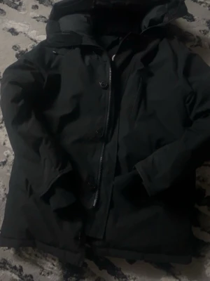 Svart parkas från Canada Goose - Säljer en svart parkas från Canada Goose med huva och klassisk logotyp på ärmen. Jackan har dragkedja och knappar framtill samt en bröstficka. Perfekt för kalla dagar och har en stilren look.