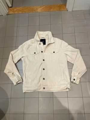 Beige overshirt med knappar - Stilren beige overshirt från med klassisk krage och knappar framtill. Två bröstfickor med lock och knapp, samt ärmar med knapp vid ärmslut. Perfekt att slänga över en hoodie eller t-shirt för en avslappnad look.