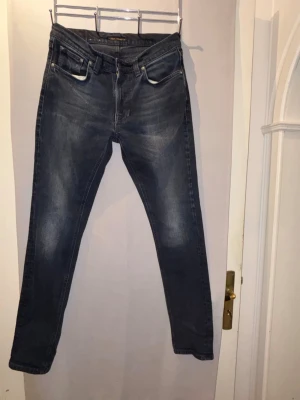Mörkblå slim jeans från Nudie W30 L32 - Snygga mörkblå jeans från Nudie med klassisk femficksdesign och diskreta slitningar framtill. Jeansen har smal passform och normal midja, perfekta till en avslappnad stil. Tillverkade i mjukt denimtyg för skön känsla.