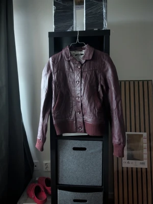 Vinröd skinnjacka med knappar - Snygg vinröd skinnjacka med knappar framtill och ribbade muddar vid ärmslut och midja. Jackan har klassisk krage och två fickor framtill. Perfekt för dig som vill ha en stilren och tidlös look.