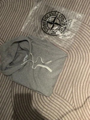 Grå hoodie från Stone Island - Snygg grå hoodie från Stone Island med klassisk huva och vita dragsnören. Tillverkad i mjukt bomullsmaterial för en skön och avslappnad look. Perfekt för dig som gillar streetwear och vill ha något stilrent och bekvämt.