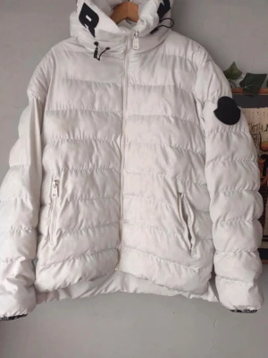 Vit pufferjacka med huva - Snygg vit pufferjacka med huva och dragkedja framtill. Jackan har två fickor med dragkedja och svarta detaljer på huvan samt ett svart märke på ena ärmen. Perfekt för kyliga dagar och ger en modern look.