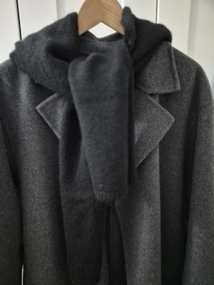 Svart stickad väst med huva - Svart stickad väst med huva och breda axelpartier. Västen har en enkel, rak form och är tillverkad i mjukt stickat material. Perfekt för lager-på-lager och ger en cool vibe till din outfit. Snygg att bära över en långärmad tröja eller jacka.