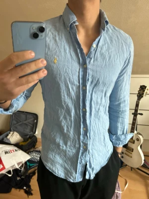 Ljusblå linen skjorta från Ralph Lauren - 100% linen Klassisk ljusblå skjorta från Ralph Lauren med button-down krage och broderad gul logga på bröstet. Skjortan har långa ärmar, vita knappar och är tillverkad i bomull. Perfekt för en clean och stilren look.