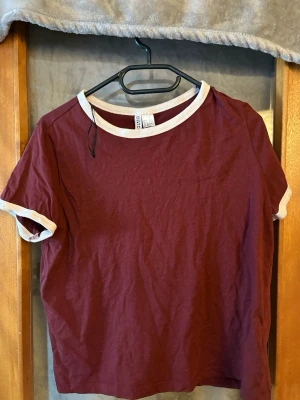 Vinröd t-shirt från H&M Divided - Säljer en vinröd t-shirt från H&M Divided med vita kontrastkanter vid halsringning och ärmslut. Klassisk modell med korta ärmar och rund hals. Perfekt till jeans eller kjol för en enkel och snygg look.