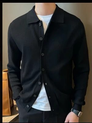 Svart ribbad cardigan med knappar - Stilren svart cardigan med ribbad struktur och knäppning framtill. Tröjan har klassisk krage och långa ärmar, perfekt att bära över en t-shirt för en clean look. Passar dig som gillar minimalistisk och tidlös stil.