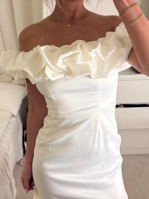Vit offshoulder klänning med volang - Superfin vit kort klänning i satin med offshoulder-modell och stor volang upptill. Sitter figurnära och har en elegant, stilren look som passar perfekt när du vill sticka ut lite extra.