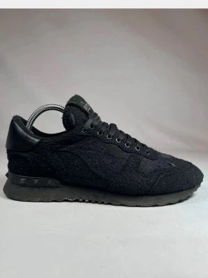 Valentino Garavani svarta sneakers - Svarta Valentino Garavani sneakers med snygg, textil och mockaliknande ovandel. Klassisk snörning och ikoniska nitar på sulan. Märkeslogga på plösen och hälparti i skinn. Perfekta för dig som gillar stilrena och exklusiva sneakers.