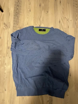 Blå tröja med prickiga armbågslappar - Snygg blå tröja från HANSON&JACOB med rund halsringning och unika armbågslappar med små gula prickar. Insidan av kragen är grön för en cool kontrast. Ena armhålan hade ett hål, men är sytt, skulle säga att det inte syns vid användning. Perfekt för dig som gillar stilrena detaljer och bekvämt material. Står XL men är som en M