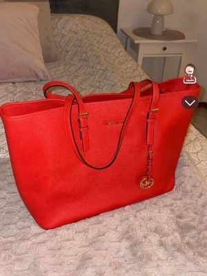 Röd handväska från Michael Kors - Snygg röd/orange handväska från Michael Kors i skinn med guldfärgade detaljer och logotyp. Väskan har dubbla handtag och en klassisk, rymlig tote-modell. Perfekt för dig som vill ha en stilren och trendig accessoar. Den är använd max en gång. Påsen/skyddet ingår. 