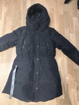 Svart dunjacka från Everest med huva - Svart lång dunjacka från Everest med huva och dragkedja framtill. Jackan har två bröstfickor med dragkedja och två stora fickor med lock. Reflexband att fälla ut från fickan. Använd max 3 ggr. 