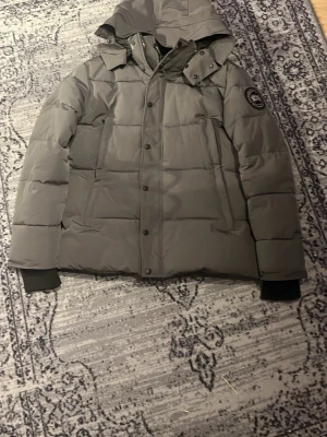 dunjacka från Canada Goose pris kan diskuteras  - Säljer en mörkgrå dunjacka från Canada Goose med huva och klassisk patch på ärmen. Jackan har knappar och dragkedja framtill, samt ribbade muddar vid ärmsluten. Perfekt för kalla vinterdagar och riktigt snygg till streetwear-looken.