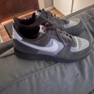 Nike Air Force 1 grå sneakers - Nike Air Force 1 sneakers i grått med ljus grå swoosh och detaljer. Ovandelen är i mocka och syntet, med perforerad tå och klassisk snörning. Yttersulan är svart och platt. Ikonisk Air Force 1-logga på hälen och tungan. Skorna är knappt använda