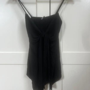 Svart linnetopp med knyt framtill - Svart ärmlös linnetopp från Gina Tricot med smala axelband och snygg knytning framtill. Toppen har en lätt asymmetrisk nederkant och är perfekt för en trendig och stilren look. Enkel att matcha med både jeans och kjol.