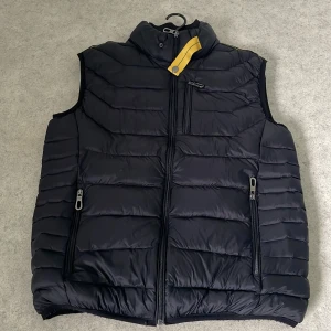 Svart dunväst från Parajumpers M - Svart dunväst från Parajumpers i storlek M. Västen har quiltad design, hög krage med gul detalj, tre dragkedjefickor framtill och klassisk logga på ryggen. Tillverkad i 100% polyester och passar perfekt för lager-på-lager under kyliga dagar.