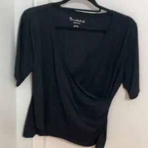 Svart omlottopp från Bondelid - Svart omlottopp från Bondelid i storlek L. Toppen har en djup v-ringning och korta ärmar, med en snygg drapering framtill. Tillverkad i mjuk och lätt nylon som ger en skön känsla. Perfekt för dig som gillar stilrena och enkla plagg med en twist.