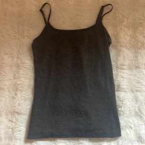 Mörkgrå linnetopp med smala axelband - En stilren mörkgrå linnetopp med smala axelband och rak passform. Toppen är ärmlös och har en enkel, klassisk design som passar perfekt till jeans eller kjol. Materialet känns mjukt och stretchigt, vilket gör den bekväm att bära hela dagen. Ny utan prislapp. 