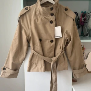 Beige croppad trenchcoat från Bershka - Snygg croppad trenchcoat från Bershka i beige bomullsmix. Jackan har dubbelknäppning, bälte i midjan, axelklaffar och stora knappar. Klassisk krage och långa ärmar med knäppning vid ärmslut. Perfekt för en trendig look i vår och höst.