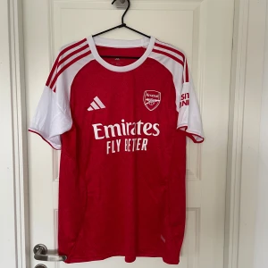 Arsenal hemmatröja Adidas röd/vit L - Säljer en officiell Arsenal hemmatröja från Adidas i storlek L. Tröjan är röd med vita ärmar och detaljer, har klubbmärke och sponsortryck framtill samt diskret mönster i tyget. Slim fit-modell i lätt och ventilerande material, perfekt för match eller träning.