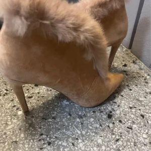 Beiga stövletter med pälsdetalj - Säljer ett par snygga beiga stövletter med spetsig tå och hög klack. Skorna är i mocka och har en fluffig pälsdetalj längs kanten som ger en lyxig vibe. Perfekta för dig som vill sticka ut med din stil.