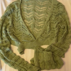 Grön omlottstickad kofta - Snygg grön stickad kofta med omlottmodell och djup v-ringning. Koftan har ett öppet, mönstrat stickat tyg och långa ärmar. Perfekt att slänga över en topp för en chill och trendig look.