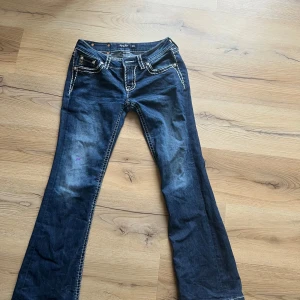 Mörkblå Miss Me jeans med bootcut - Snygga mörkblå jeans från Miss Me med tydliga kontrastsömmar och broderade bakfickor. Bootcut-modell med klassisk femficksdesign och lila detalj på ena benet. Jeansen har låg midja och är tillverkade i stretchig denim för skön passform. En liten lila fläck som inte syns på håll