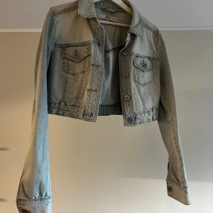 Ljusblå croppad jeansjacka JSFN - Snygg ljusblå croppad jeansjacka från JSFN med klassisk krage, två bröstfickor med knappar och långa ärmar. Jackan har en stilren design med silverfärgade knappar och sömdetaljer. Perfekt för dig som vill ha en trendig och avslappnad look.