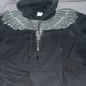 Svart hoodie Marcelo Burlon med vingar - Svart hoodie från Marcelo Burlon County of Milan med ikoniskt vingtryck över axlar och bröst. Tröjan har huva med snörning, känguruficka och ribbade muddar. Grym streetwear-vibe och perfekt för dig som gillar unika detaljer. Priset kan diskuteras 