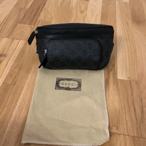 Svart Gucci axelväska med GG-mönster - Snygg svart axelväska från Gucci med klassiskt GG-mönster i mörk ton. Väskan har justerbart band, dragkedja upptill och extra fack framtill. Tillverkad i skinn med detaljer i silverfärgad metall. Perfekt för att bära det viktigaste med stil.