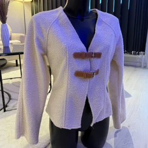 Beige teddyjacka med spännen - Supermysig beige teddyjacka med två snygga bruna spännen framtill. Jackan har lång ärm, rak passform och är tillverkad i ett fluffigt teddy-material som ger en cool och trendig look. Perfekt för dig som vill ha något både stilrent och unikt. Passar perfekt som oversize modell.