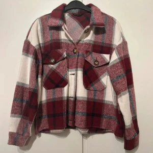 Rutig höstjacka i vinrött och vitt - Höstjacka med stora rutor i vinrött, vitt och grått. Jackan har två bröstfickor med knappar, klassisk krage och långa ärmar. Perfekt lager-på-lager-plagg för höst. Mjuk och skön känsla, stängs med knappar framtill. Fint skick!