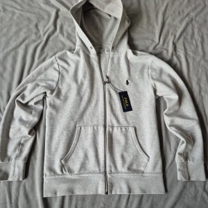 Grå hoodie från Polo Ralph Lauren - Snygg ljusgrå hoodie från Polo Ralph Lauren med dragkedja och klassisk broderad logga på bröstet. Tröjan har huva, känguruficka och är tillverkad i mjuk bomullsmix. Perfekt för chill dagar eller när du vill ha en clean look.