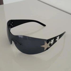 Svarta y2k solglasögon med silver stjärnor  - Säljer ett par svarta solglasögon med futuristisk shield-form och spegelblåa linser, väldigt 2000-tal. Sidorna är dekorerade med stora silvriga stjärnor som ger en riktigt cool vibe. Perfekta för dig som vill sticka ut och addera edge till din stil.