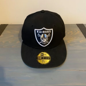 Svart Raiders keps - Säljer en svart 59FIFTY keps från New Era med Raiders-logga broderad framtill och NFL-märke baktill. Klassisk flat brim och stilren design. Kepsen är i bomull och har broderade detaljer på sidorna. Hör av er vid minsta fundering!