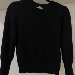 Svart glittrig tröja från H&M - Svart tröja från H&M med diskret glittrig effekt. Tröjan har rund halsringning, långa ärmar med lätt puff vid axlarna och ribbade muddar. Fint skick!