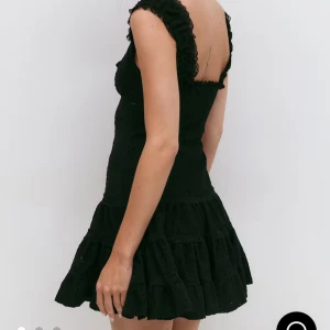 Svart broderad kortklänning från ginatricot  - Supersöt svart klänning från gina i storlek M. Den har broderade detaljer, volangkant vid axlarna och en liten knytning framtill. Klänningen är ärmlös och har en kort, voluminös kjol med spets och mönstrad struktur. Perfekt för dig som gillar romantisk och trendig stil. 