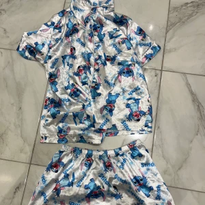 Stitch pyjamasshorts i satin - Säljer ett par silvriga pyjamasshorts med Stitch-motiv från Lilo & Stitch. Shortsen är i glansigt satinmaterial med elastisk midja och har ett färgglatt tryck i blått, rosa och rött. Perfekta för dig som älskar Disney och vill ha något bekvämt och lekfullt att chilla i.