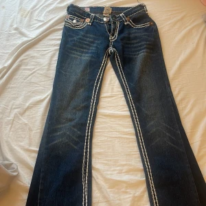 True Religion jeans med vita sömmar - Byxorna är väldigt små i storleken! Mörkblå jeans från True Religion med tydliga vita kontrastsömmar och stora fickor bak med dekorativa knappar. Klassisk bootcut-modell i jeansmaterial. Snygga detaljer och ikonisk logga på bakfickan. 
