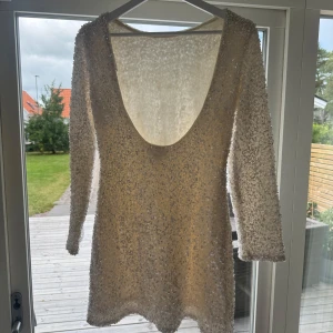 Paljettklänning med öppen rygg - Säljer en kort, ljus paljettklänning med långärmade ärmar och djup öppen rygg. Klänningen är täckt av vita paljetter som ger ett glittrigt och festligt intryck. Materialet känns mjukt och har en lyxig känsla, perfekt för att sticka ut. Passformen är normal och klänningen har en snygg siluett.