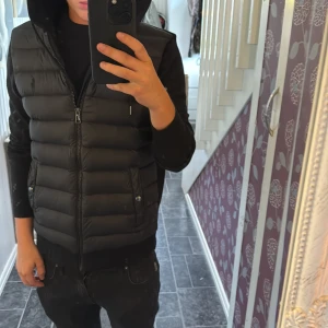 Svart dunväst från Polo Ralph Lauren - Svart dunväst från Polo Ralph Lauren Performance med huva och snörning. Västen har quiltad framsida, två fickor med tryckknappar och dragkedja framtill. Baksidan är i mjuk bomull och huvan har snygga detaljer. Perfekt för lager-på-lager och streetwear. Liten defekt på insidan vid dragkedjan. Annars mycket fint skick sitter bra på mig som är 170