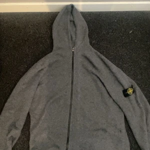 Stone Island merinoull - Grå hoodie från Stone Island  med dragkedja framtill och klassisk logga på ärmen. Tröjan har huva och är tillverkad i mjukt stickat material. Perfekt för dig som gillar merinoull och vill ha en stilren look.
