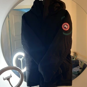 Svart dunjacka från Canada Goose S - Säljer en svart dunjacka från Canada Goose i storlek S. Jackan har huva, klassisk patch på ärmen och stilren design. Perfekt för kalla dagar med sin varma och fluffiga känsla. Jackan har dragkedja och praktiska fickor framtill.