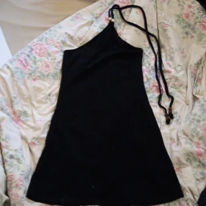 Svart halterneckklänning divided H&M S - Svart kort klänning från divided H&M i halterneckmodell med smala band och pärldetalj vid halsen. Sitter tight och har en enkel, stilren look. Perfekt för dig som gillar minimalistisk stil och vill ha något snyggt och bekvämt.