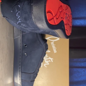 Mörkblå Christian Louboutin sneakers - Säljer ett par mörkblå sneakers från Christian Louboutin i lyxig mocka med signaturbroderi på hälen och ikonisk röd sula. Snygg, stilren design med platt sula och snörning. Perfekt för dig som vill sticka ut med exklusiva detaljer och premiumkänsla.