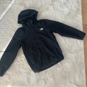 Svart regnjacka The North Face DryVent - Svart regnjacka från The North Face med DryVent-teknologi. Jackan har huva, dragkedja framtill och meshfoder på insidan. Perfekt för blöta dagar och har en clean, sportig look med logga på bröstet.