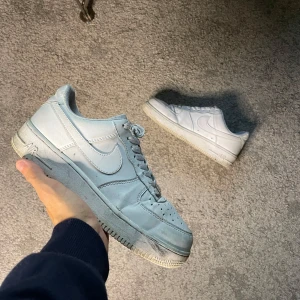 Nike Air Force 1 vita sneakers - Klassiska Nike Air Force 1 sneakers i helvitt skinn med perforerad tå, snörning och platt sula. Ikonisk siluett med rund tå och diskret Nike-logga på sidan. Perfekta för en clean och stilren look.