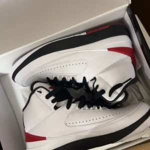 Nike Air Jordan 2 vita och röda - Säljer ett par Nike Air Jordan 2 sneakers i vitt skinn med svarta och röda detaljer. Klassisk high-top siluett med svart snörning och perforerade paneler på sidorna. Snygg kontrast mellan den vita ovandelen och den svarta sulan, med röda accenter vid hälen.