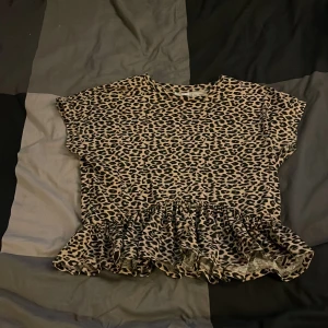 Leopardmönstrad topp med volang Kappahl - Snygg topp från Kappahl i leopardmönster med volangkant nedtill. Toppen har korta ärmar och är i mjuk, stretchig bomull. Perfekt för dig som vill sticka ut med ett djurmönster och gillar trendiga detaljer.