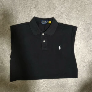 Svart pikétröja från Polo Ralph Lauren - Klassisk svart pikétröja från Polo Ralph Lauren med vit broderad logga på bröstet. Helt ny, aldrig använd.Tröjan har korta ärmar, krage och två knappar framtill. Tillverkad i mjuk bomull som känns skön mot huden. Perfekt för en clean och stilren look.